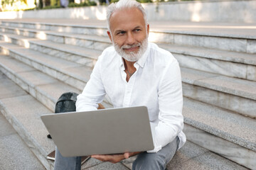 Smiling man in stylish light outfit poses with laptop. Joyful guy in white trendy shirt with long sleeve sits on stairs..