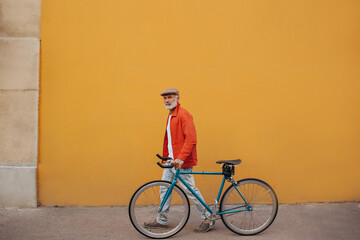 Full length shot of man in cap and orange jacket walking with bicycle. Bright adult in jeans and...