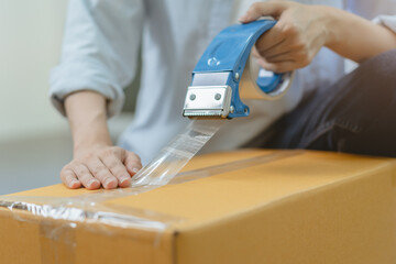 Focus hand of owner business asian woman, female holding packing box by tape machine, sealing cardboard indoors. Business owner in warehouse, Moving or preparing in new house, apartment or relocation.