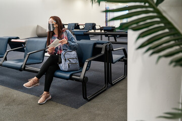 latina female tourist with face mask and face shield checking her flight tickets and sitting in a socially distanced chair at the airport during the coronavirus pandemic or covid19 virus
