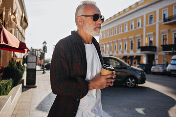 Tattooed man in plaid shirt and sunglasses holds tea cup. Attractive adult guy in white T-shirt and glasses posing outdoors..