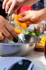 manos exprimiendo el jugo de un limón amarillo en un bol metálico, con mesa llena de diferentes utensilios 