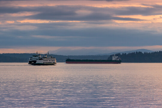 As The Olympic Mountains Rise Into An Overcast Sky At Dusk, A Washington State Ferry And A Freighter Container Ship Pass On Puget Sound In The Pacific Northwest.