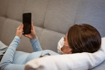 tired caucasian woman in casual clothes in a protective mask on the couch with a phone