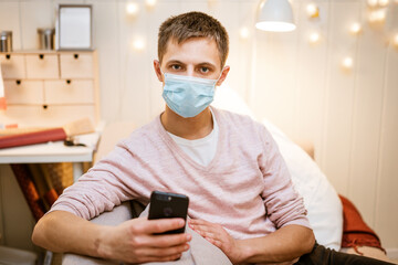 young caucasian man sitting on a sofa in casual clothes, wearing a protective medical mask with a phone in his hand