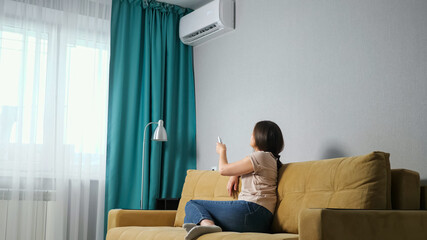 side view of a brunette woman adjusts the air conditioner while sitting on the sofa. - obrazy, fototapety, plakaty