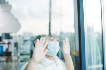 Cute asian young boy wearing medical mask, standing idle in front of close window, looking through window,