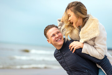 Happy man giving piggyback ride to his woman and laughing at beach. Smiling guy in love carrying on...