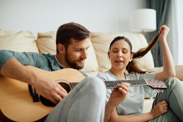 Obraz premium Young woman sings while her husband is playing acoustic guitar at home.