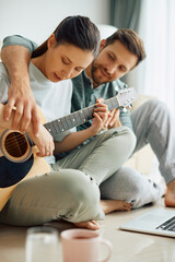 Young woman learns to play acoustic guitar with help of her husband at home.