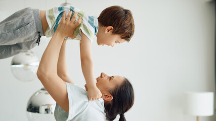 Playful mother holds her small son high up and has fun with him at home.