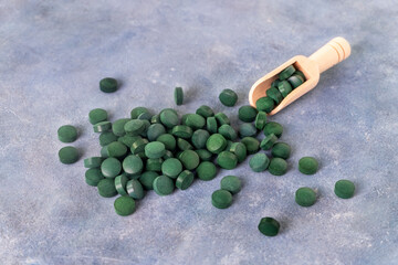 Green pills of spirulina or chlorella on a gray-blue plaster background in a glass bowl and in a wooden spoon. Healthy superfood diet and detox nutrition concept.