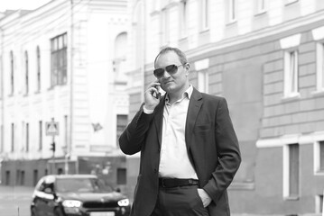 Black and white portrait of a young caucasian man in a business suit and sunglasses holds a smartphone for business negotiations. Work, business. Copy space on the left