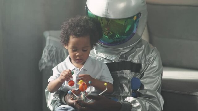 African father and son play in the living room at home, man in an astronaut costume sitting on the floor with her son, boy playing with a toy model of the solar system, astronomy.