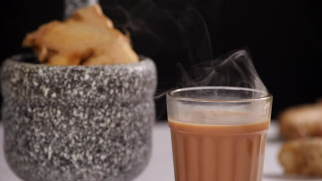 Smoke coming out of a boiling hot aromatic ginger tea served in a clear glass - Indian Chai. Closeup shot of a piece of Adrak kept in a mortar pestle on a white table against a dark background