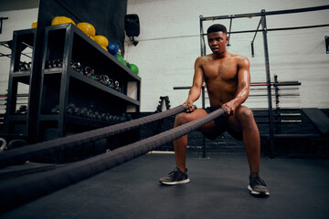 Fototapeta na wymiar Young African American male exercising inside a gym. Topless male personal trainer using rope to strengthen his arms. High quality photo