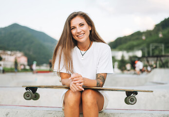 Slim young woman with long blonde hair in light sports clothes with longboard in outdoor skatepark...
