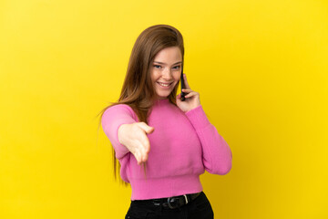 Teenager girl using mobile phone over isolated yellow background shaking hands for closing a good...