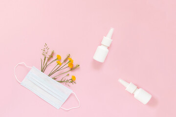A set of wild flowers and herbs under a disposable protective mask and a mock-up of nasal spray on a light pink background. The concept of means of protection and treatment of seasonal allergies.