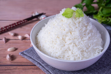 Steamed organic white rice in a white ceramic bowl with chopsticks and apron and green coriander stained on a brown wooden table.