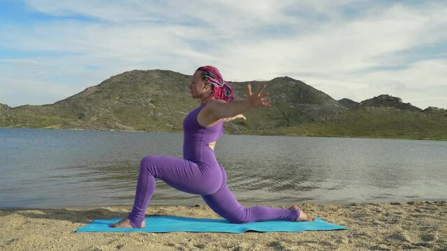 Young woman with dreadlocks doing yoga poses, having training on the lake shore