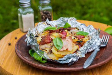 Baked sliced potatoes with pork belly and onions in foil on a brown clay plate on a light wooden background. Potato dishes.