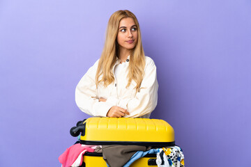 Young Uruguayan blonde woman with a suitcase full of clothes over isolated purple wall looking to the side