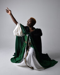 Full length portrait of pretty African woman wearing long green medieval fantasy gown, kneeling pose on a light grey studio background. © faestock