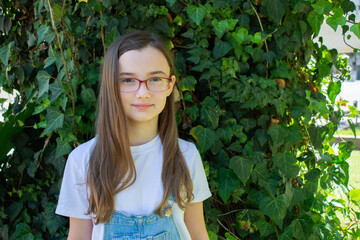 A girl with long brown hair. A girl in a white T-shirt and denim overalls looks at us. The girl on the background of the wall covered with ivy.