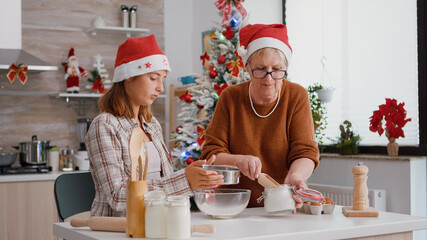 Senior woman putting flour in metalic siev while granddaughter sift ingredient in bowl preparing xmas traditional cookie. Family enjoying christmas holiday together in decorated kitchen
