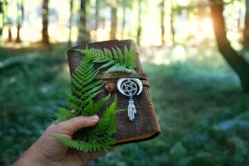 Magic witch book with amulet and fern leaves in hand on natural forest background. Esoteric Ritual,...