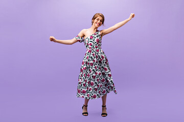 Fototapeta na wymiar Cheerful girl in floral print dress dances on purple background. Lovely beautiful young woman in fashionable summer outfit and black heels has fan..