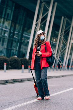 Portrait of a traveler woman in a mask walking with an orange suitcase near an airport. Young fashionable woman in a blue jeans and jacket