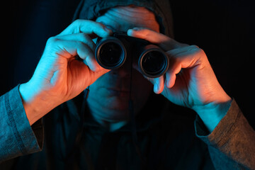middle-aged man, stalker in hood looks through black binoculars in dark, peeps, illegally tracks...