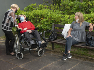Closeup of a sick older man wearing a mask in a wheelchair next to two women helpers in a park