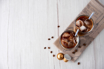 Iced coffee latte in a tall glass with cream and beans, golden capsules, straws on light background. Cold tasty summer refreshment beverage concept. Selective focus, copyspace. Flat lay, top view.