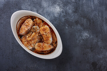 Baked white sea fish macrourus in marinade of olive oil, rosemary, spices in white pan on dark textured background. Top view, copy space