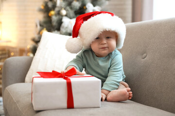 Obraz na płótnie Canvas Cute baby in Santa hat with Christmas gift sitting on sofa at home