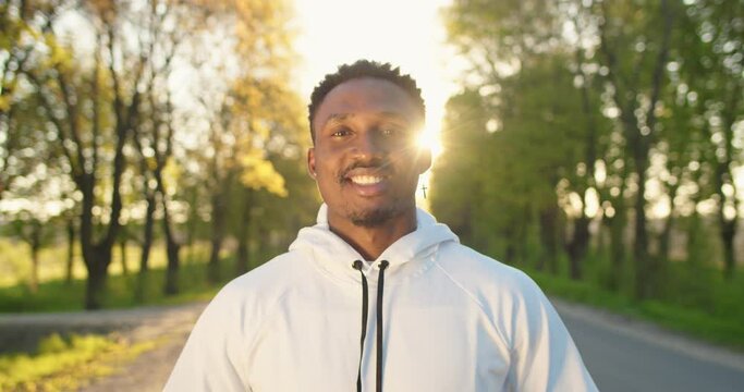 Close up of joyful handsome young African American man looking at camera and smiling while standing alone on street in town in the morning on sunrise. Happy sportsman at outdoor workout