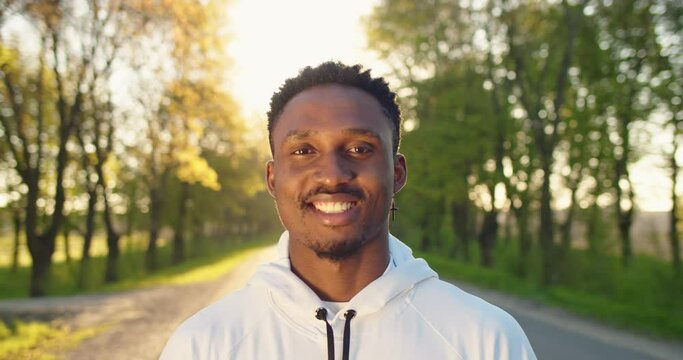 Close up portrait of joyful positive handsome African American guy in good mood smiling at camera standing on street in sun lights. Happy young male with smile on face, outdoor workout, sport concept