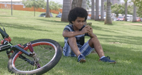 The young boy sits on the grass in the garden and using smartphone.