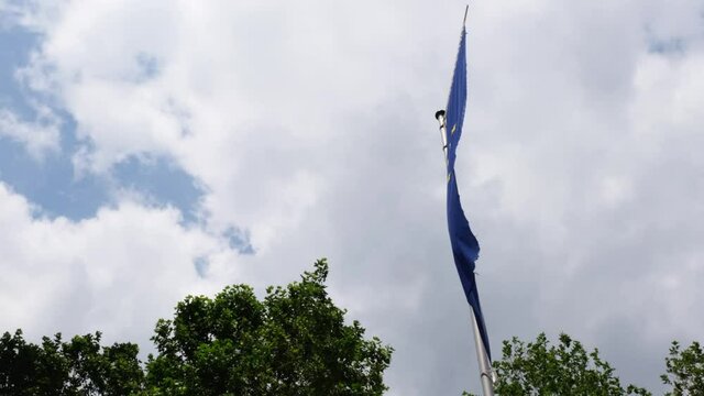 Waving European Union flag against the cloudy sky. Blue flag with yellow stars fluttering in the afternoon breeze. EU flag and flag poole blowing in the wind. Windy summer day in Düsseldorf, Germany.