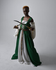 Full length portrait of pretty African woman wearing long green medieval fantasy gown holding  sword, standing action pose on a light grey studio background.