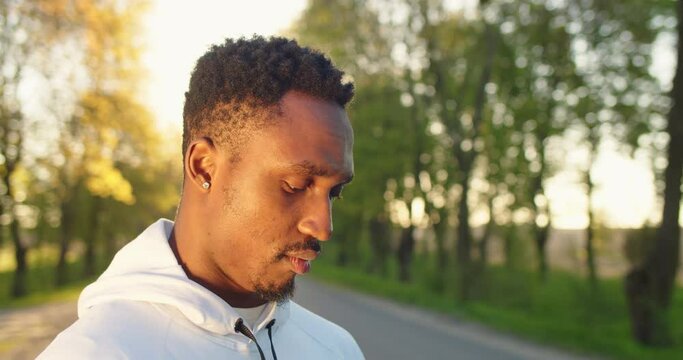 Close up portrait of joyful handsome African American guy standing alone on street in sun lights putting wireless earphones preparing for workout outdoor. Young male athlete listening to music outside