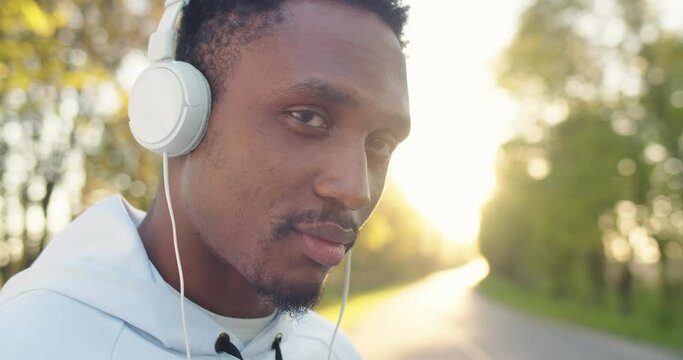 Close up shot of cheerful smiling young handsome African American guy sportsman standing outdoors putting headphones and looking at camera listening to music before workout. Leisure concept