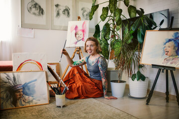 Charming caucasian red-haired woman artist with tattoos in home studio, light photography of...