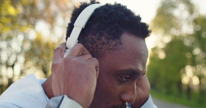 Close up of young handsome African American guy standing alone on street listening to music in white headphones, outdoor workout. Male athlete, training outside, sport, recreation, leisure concept