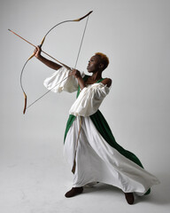 Full length portrait of pretty African woman wearing long green medieval fantasy gown holding bow and arrow, standing action pose on a light grey studio background.