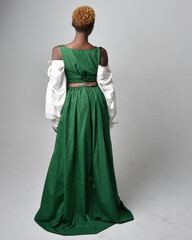 Full length portrait of pretty African woman wearing long green medieval fantasy gown, standing pose facing backwards on a light grey studio background.