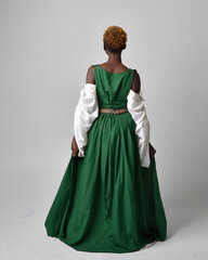 Full length portrait of pretty African woman wearing long green medieval fantasy gown, standing pose facing backwards on a light grey studio background.